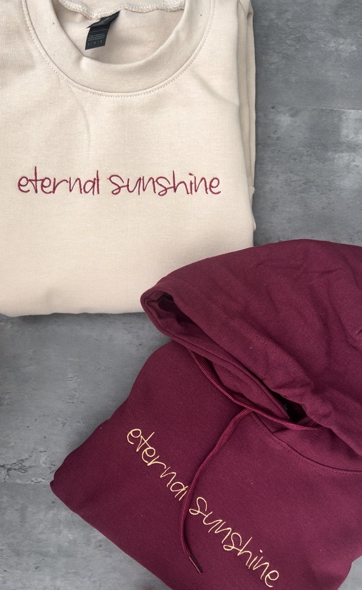 Ariana grandes eternal sunshine album font and text embroidered onto a maroon sweatshirt in a gold colour and also embroidered onto a beige sweatshirt in a maroon colour