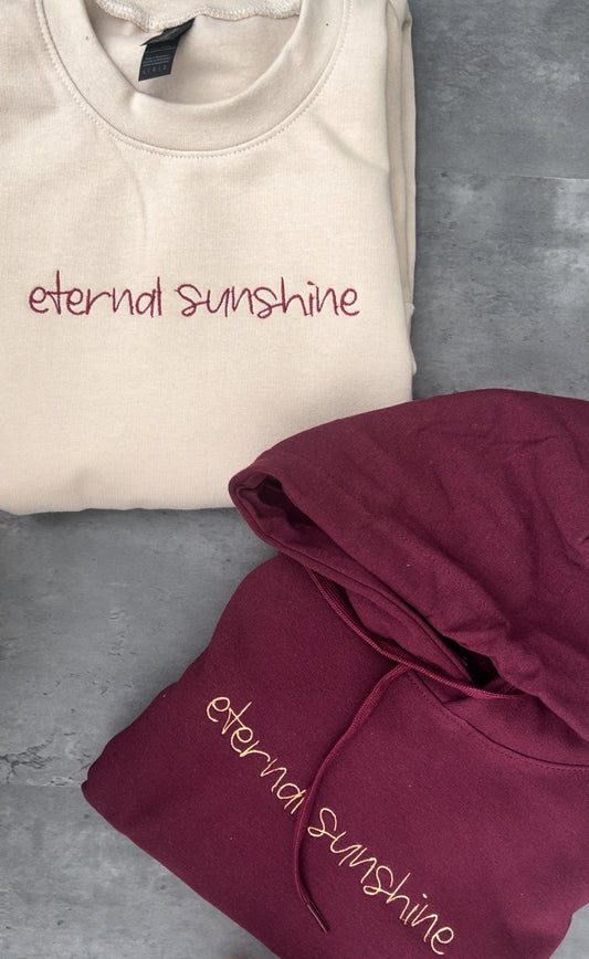Ariana grandes eternal sunshine album font and text embroidered onto a maroon sweatshirt in a gold colour and also embroidered onto a beige sweatshirt in a maroon colour