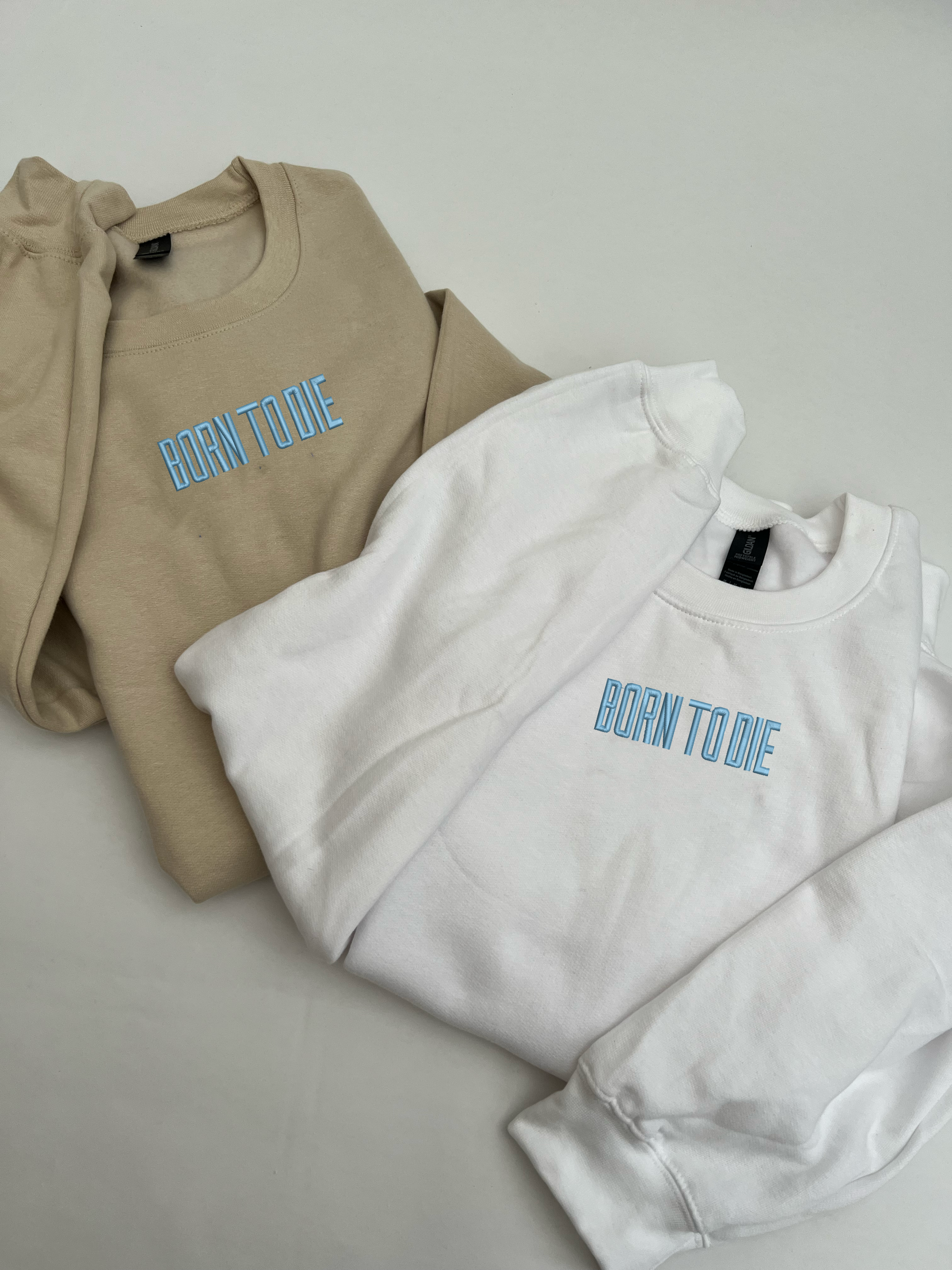 two sweatshirts folded, one sand colour and the other a white colour, lana del Reys born to die is embroidered on the centre of the chest, in the colour and font as the title appears on the album cover